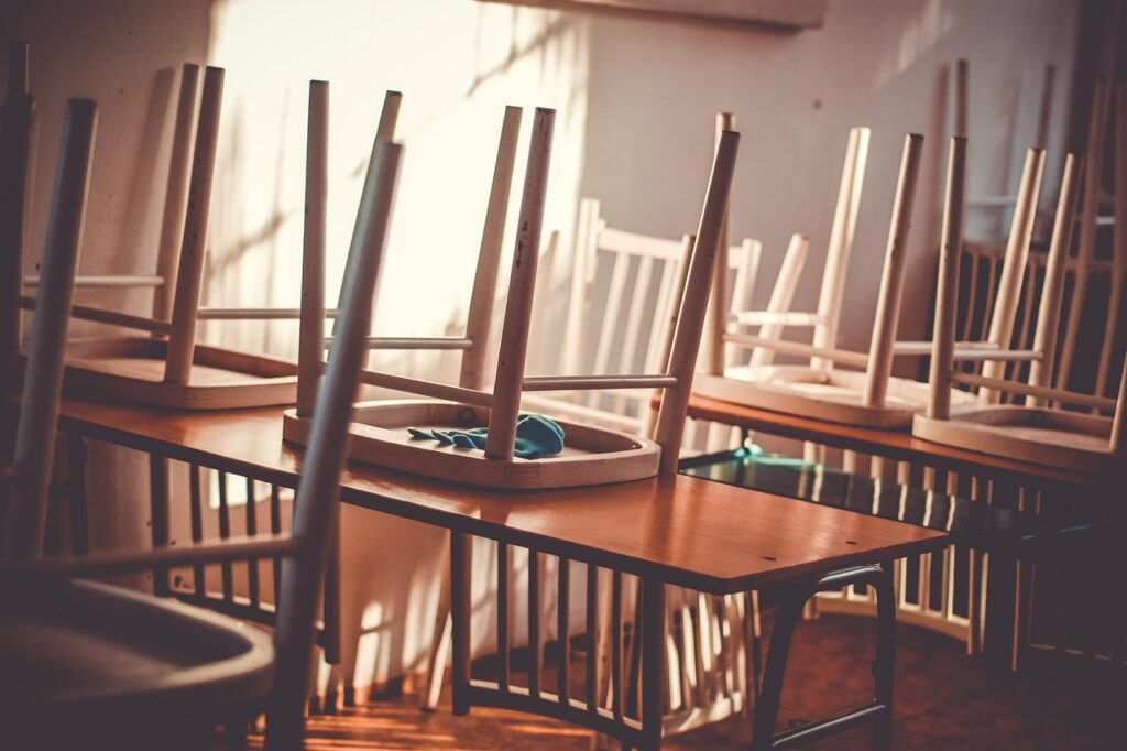 Cadeiras viradas sobre as mesas em uma sala de aula vazia, com luz de janela ao fundo