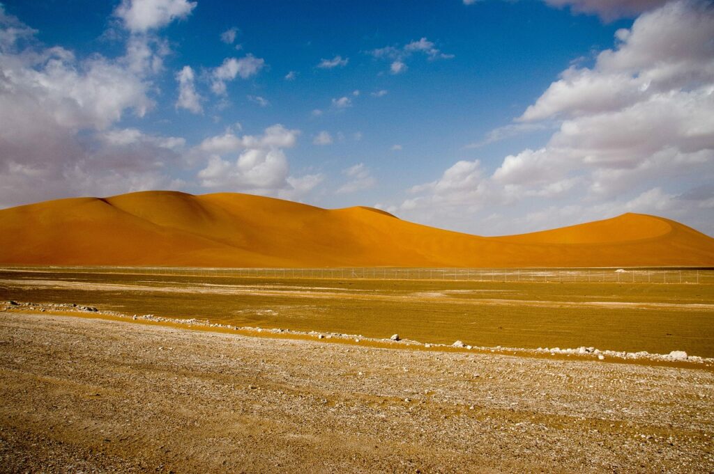 Dunas de areia alaranjadas e planície de cascalho em deserto sob céu azul com nuvens