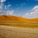 Dunas de areia alaranjadas e planície de cascalho em deserto sob céu azul com nuvens