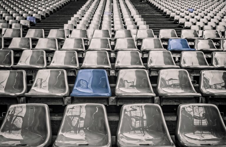 Arquibancada vazia de estádio, com fileiras de assentos cinza e um único assento azul em destaque