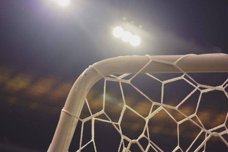 Trave de futebol com rede branca sob refletores acesos à noite, com fundo escuro e desfocado