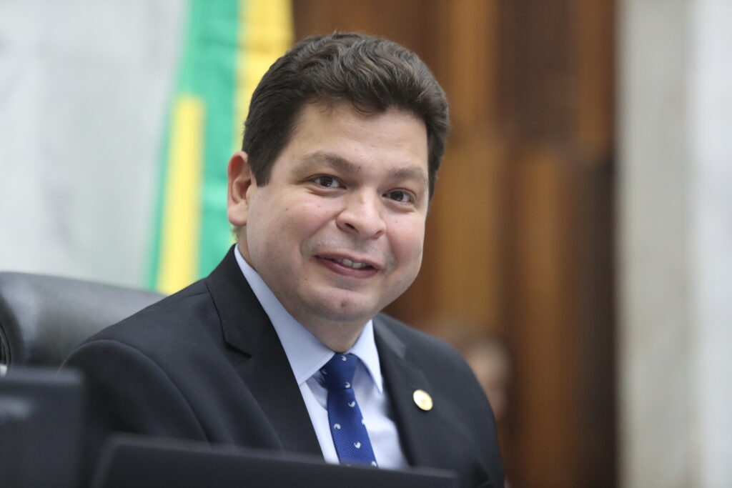 Homem de terno escuro e gravata azul sorri em ambiente institucional, com a bandeira do Paraná ao fundo