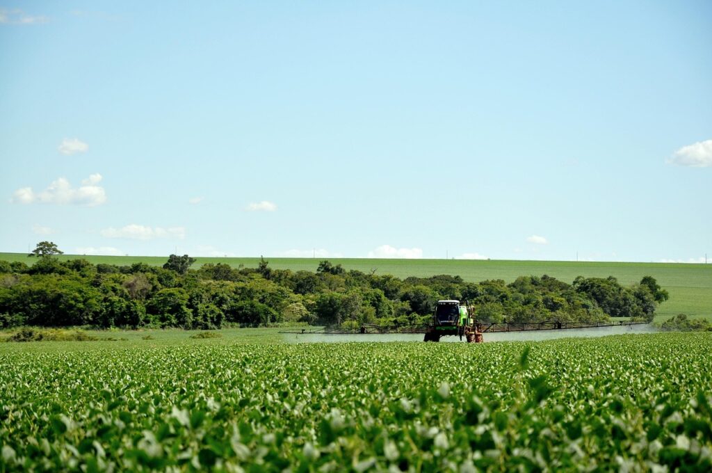 Pulverizador aplicando produto em lavoura de soja verde, com vegetação e céu azul ao fundo