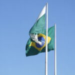 Bandeira do Brasil e outra bandeira com brasão tremulando em dois mastros contra céu azul