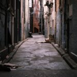 Beco urbano com papelões e lixo espalhados no chão entre muros escuros e grafitados